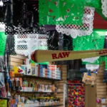 Green and white papel picado decorations with oversized RAW joint prop hanging above product aisles at Fox Plaza Smoke Shop