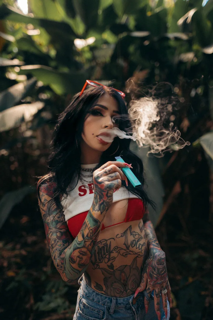 Un adulto exhalando una nube de vapor mientras sostiene un vape desechable color verde azulado al aire libre en El Paso Smoke Shops