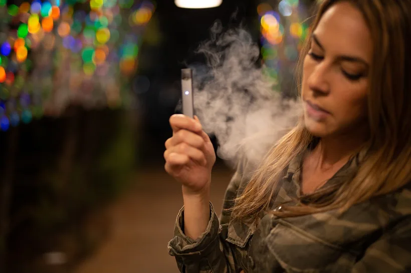 Un adulto sosteniendo un pod vape delgado y exhalando vapor bajo luces colgantes de noche en El Paso Smoke Shops