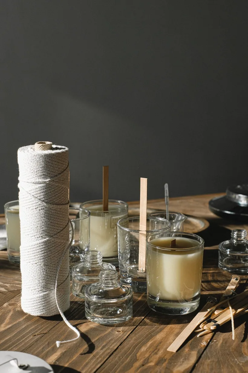 Empty candle jars filled with poured wax, a spool of cotton wick, and wooden wick-centering sticks on a wood table at El Paso Smoke Shops