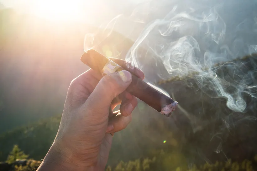 Una mano sosteniendo un puro tamaño robusto recién encendido con un hilo de humo en El Paso Smoke Shops